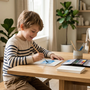 A 7-year-old boy practicing watercolor painting at a tidy wooden table in a bright, modern living room.