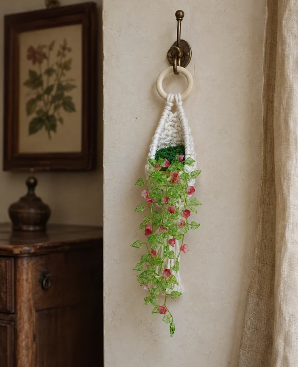 A crocheted hanging planter with a white ring handle, green stem, and pink flowers, hanging on a wall hook in a rustic room setting.