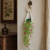A crocheted hanging planter with a white ring handle, green stem, and pink flowers, hanging on a wall hook in a rustic room setting.