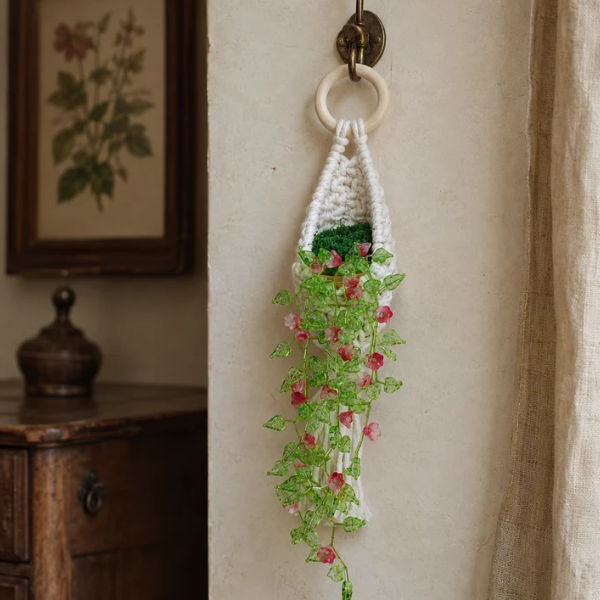 A crocheted hanging planter with a white ring handle, green stem, and pink flowers, hanging on a wall hook in a rustic room setting.
