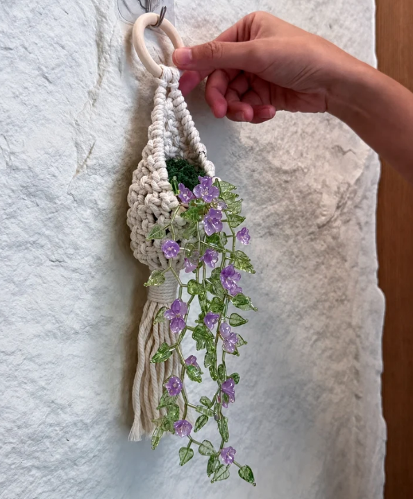 A hand holding a cream macrame plant hanger decorated with faux green leaves and purple flowers, hanging against a textured white wall.