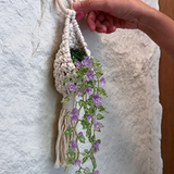 A hand holding a cream macrame plant hanger decorated with faux green leaves and purple flowers, hanging against a textured white wall.