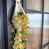 A cream macrame plant hanger with a light wood ring, holding a faux green ivy vine with small orange flowers, hanging on a dark paneled door.