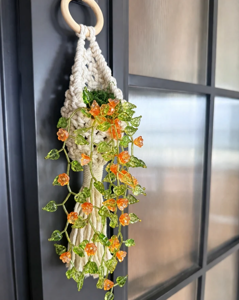 A cream macrame plant hanger with a light wood ring, holding a faux green ivy vine with small orange flowers, hanging on a dark paneled door.