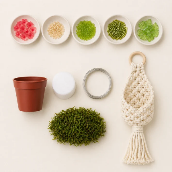 A flat-lay of a macrame plant hanger kit on a warm off-white background, including a cream-colored woven macrame hanger with a wooden ring and tassel, a green faux moss ball, a terracotta-colored plastic pot, a white foam insert, a metal wire loop, and five small bowls containing colorful beads.