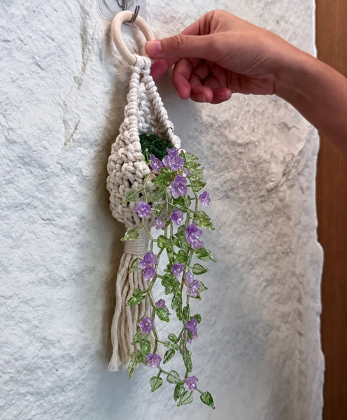 A hand holding a cream macrame plant hanger decorated with faux green leaves and purple flowers, hanging against a textured white wall.