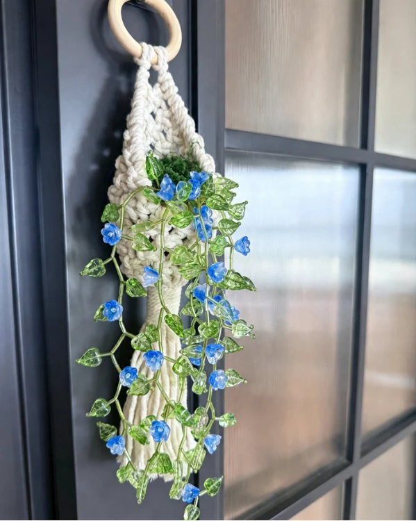 A macrame wall hanging with a light wood ring, cream cord, and green leafy vines adorned with small blue flowers, hanging on a dark paneled door.