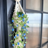 A macrame wall hanging with a light wood ring, cream cord, and green leafy vines adorned with small blue flowers, hanging on a dark paneled door.