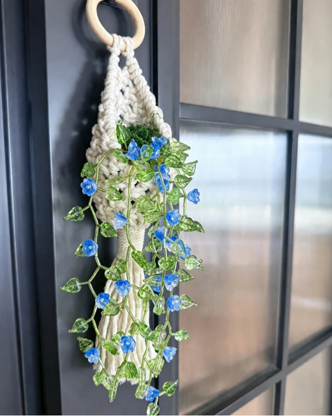 A macrame wall hanging with a light wood ring, cream cord, and green leafy vines adorned with small blue flowers, hanging on a dark paneled door.