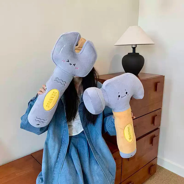 Person holding large plush wrench and hammer in a room with a wooden dresser and lamp.