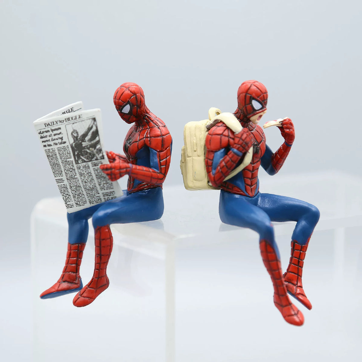 Two Spider-Man action figures sitting and reading a newspaper on a light gray background