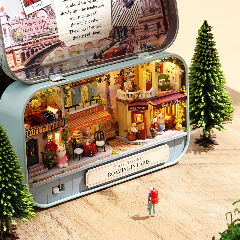 Model of a miniature town inside a tin with a Christmas tree and person figurine on a wooden floor.