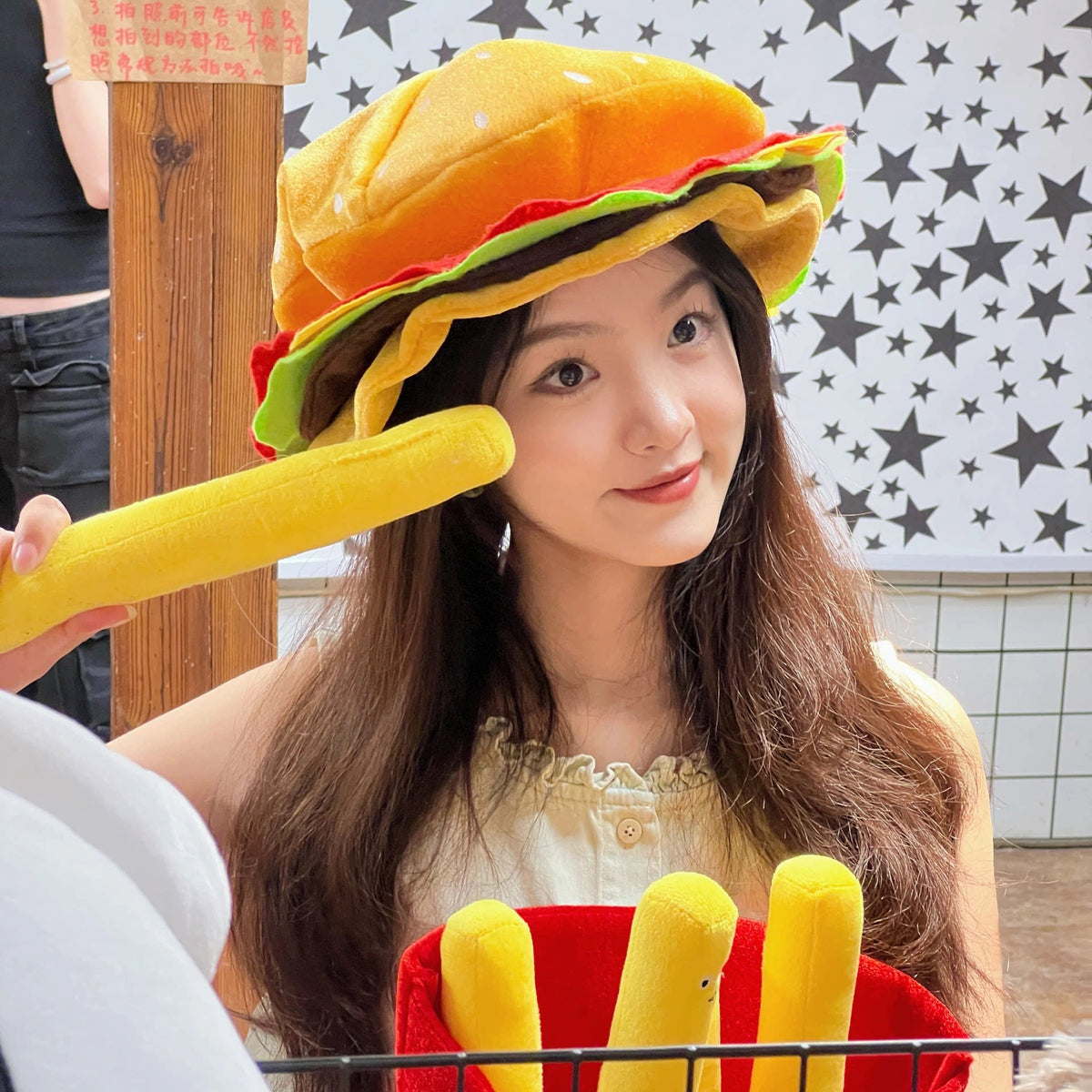 Person wearing a yellow hat and holding a toy resembling a hamburger and fries against a star-patterned wall.