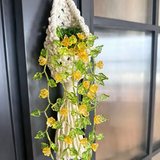A macrame wall hanging with a woven cream cord base, decorated with green variegated ivy leaves and small yellow flowers, hanging against a dark window frame.