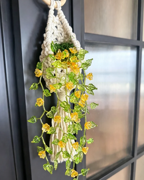 A macrame wall hanging with a woven cream cord base, decorated with green variegated ivy leaves and small yellow flowers, hanging against a dark window frame.